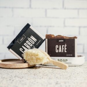 A close-up of artisanal coffee and charcoal soaps with a bath brush against a white brick wall.